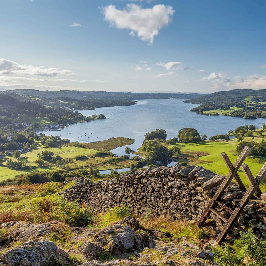 View of a serene lake surrounded by lush green hills, sailboats, and a stone wall with wooden steps in the foreground. - Home Instead