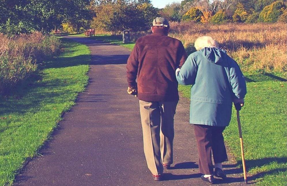 An elderly couple walks hand in hand down a paved path in a park, with the woman using a cane for support. - Home Instead