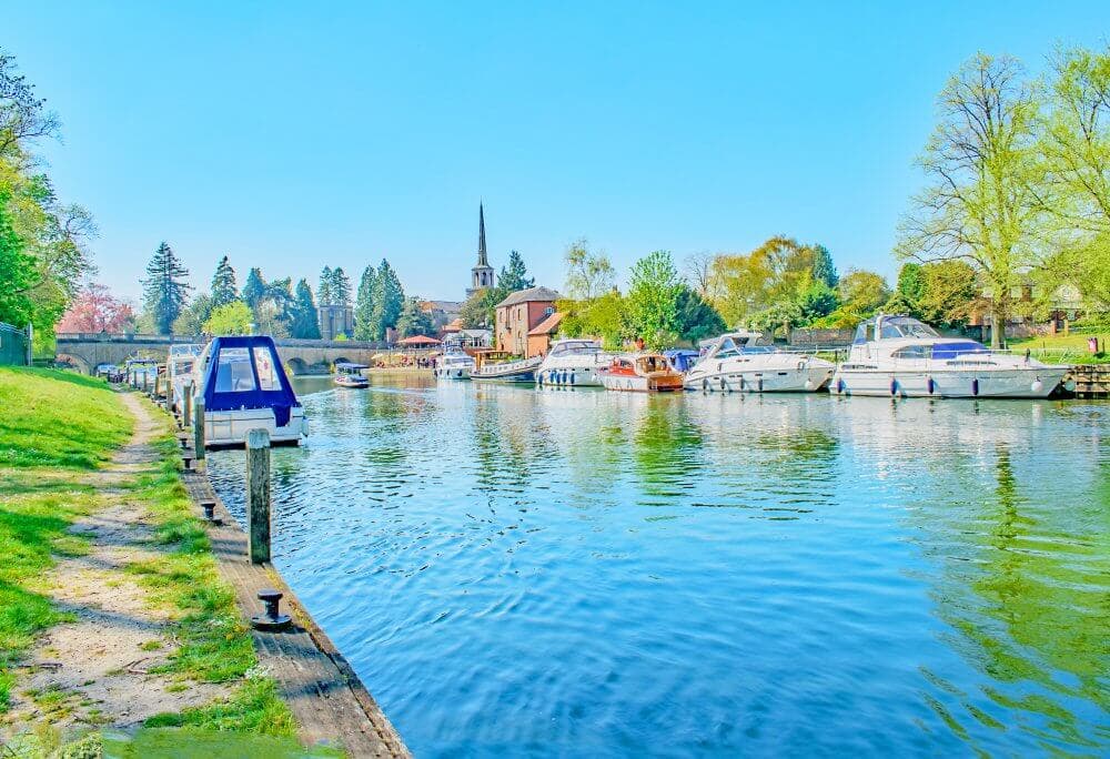 Boats docked along a serene riverside with a church and greenery in the background on a bright, sunny day. - Home Instead