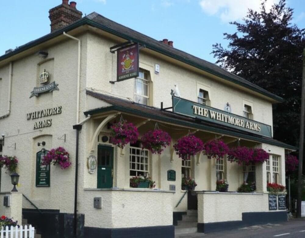 A cozy, cream-colored pub named "The Whitmore Arms," adorned with hanging flower baskets and outdoor seating. - Home Instead