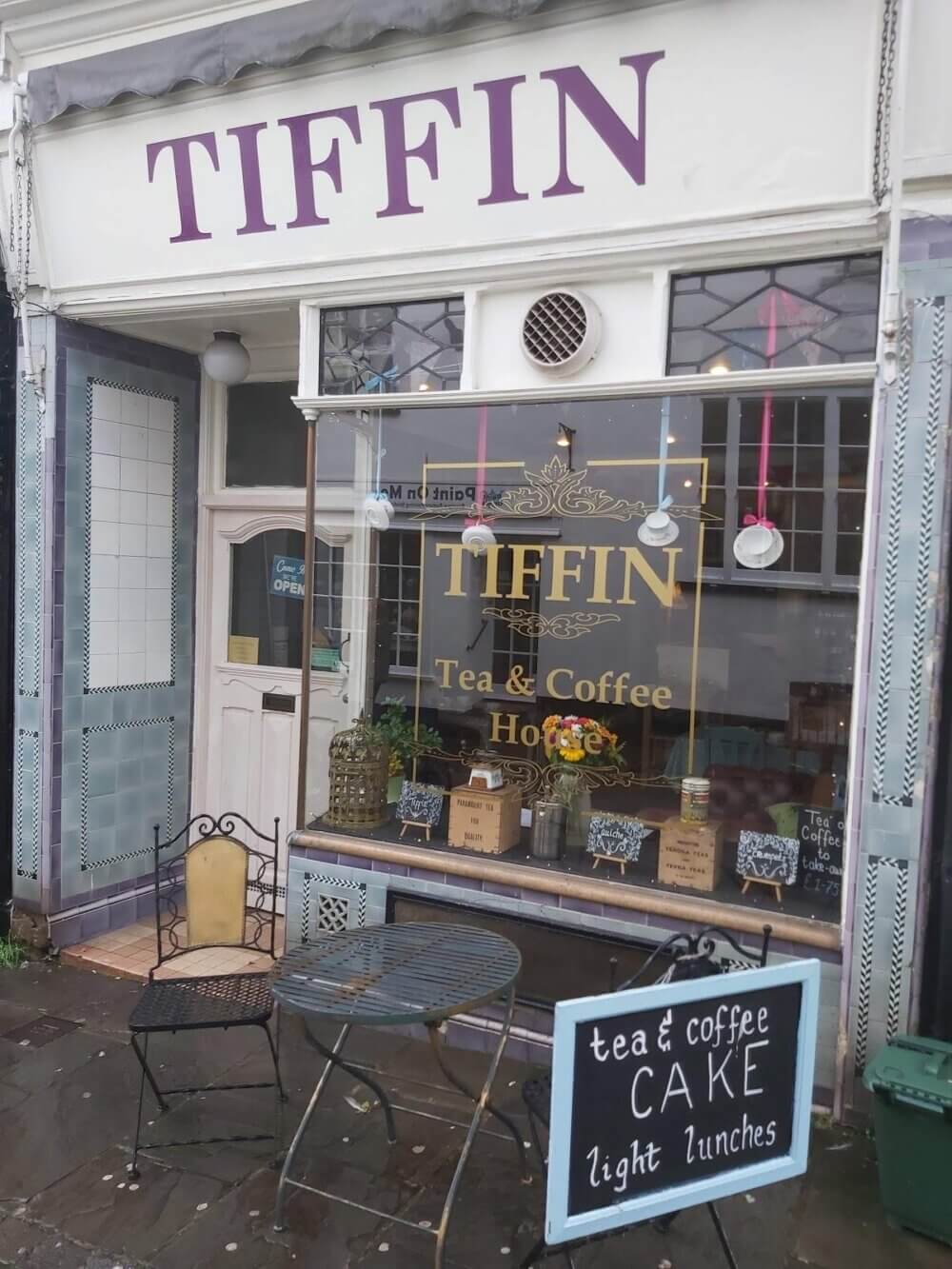 Exterior of the Tiffin Tea & Coffee House with an outdoor seating area and sign advertising tea, coffee, and cake. - Home Instead