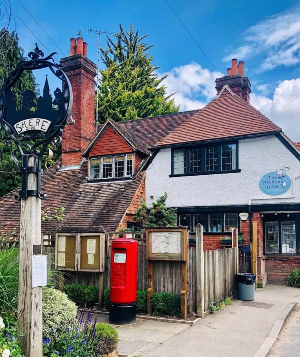 A quaint, old brick and white house with a red post box in front, a wooden fence, and a sign reading "Shere" on the street. - Home Instead