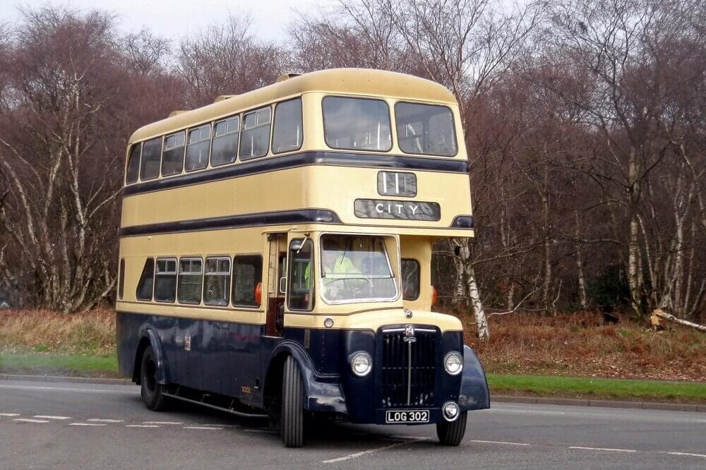 A vintage double-decker bus, marked "11 CITY," drives on a road surrounded by trees without leaves. - Home Instead