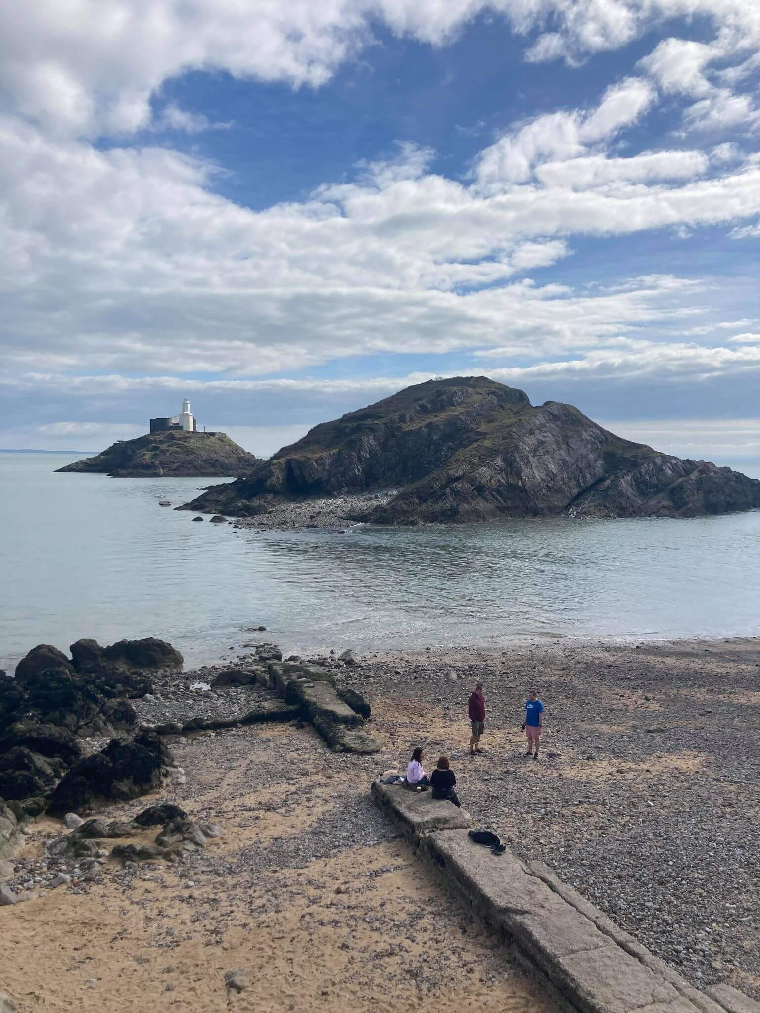 A group of people stand on a pebble beach overlooking a rocky island with a lighthouse under a cloudy sky. - Home Instead