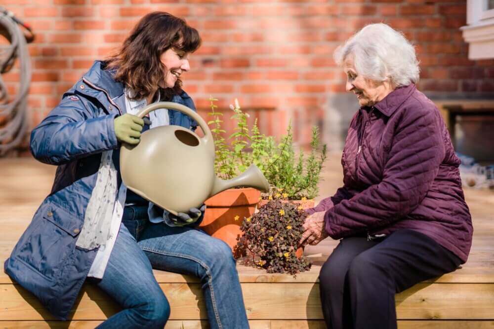 Two women gardening together on a wooden deck, one watering plants while the other tends to a pot of flowers. - Home Instead