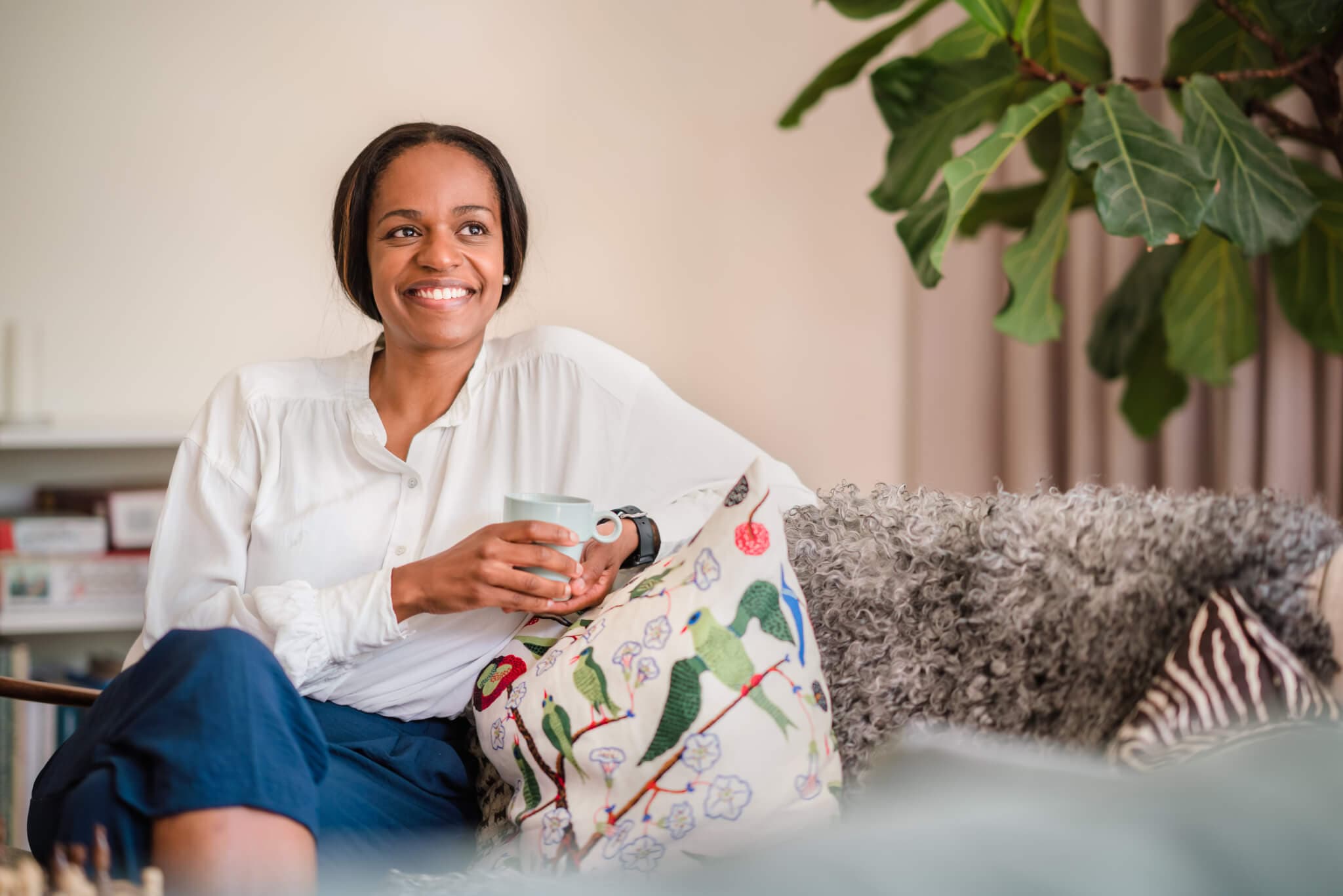 A smiling woman sits on a couch holding a mug, with a decorative pillow and a large plant in the background. - Home Instead