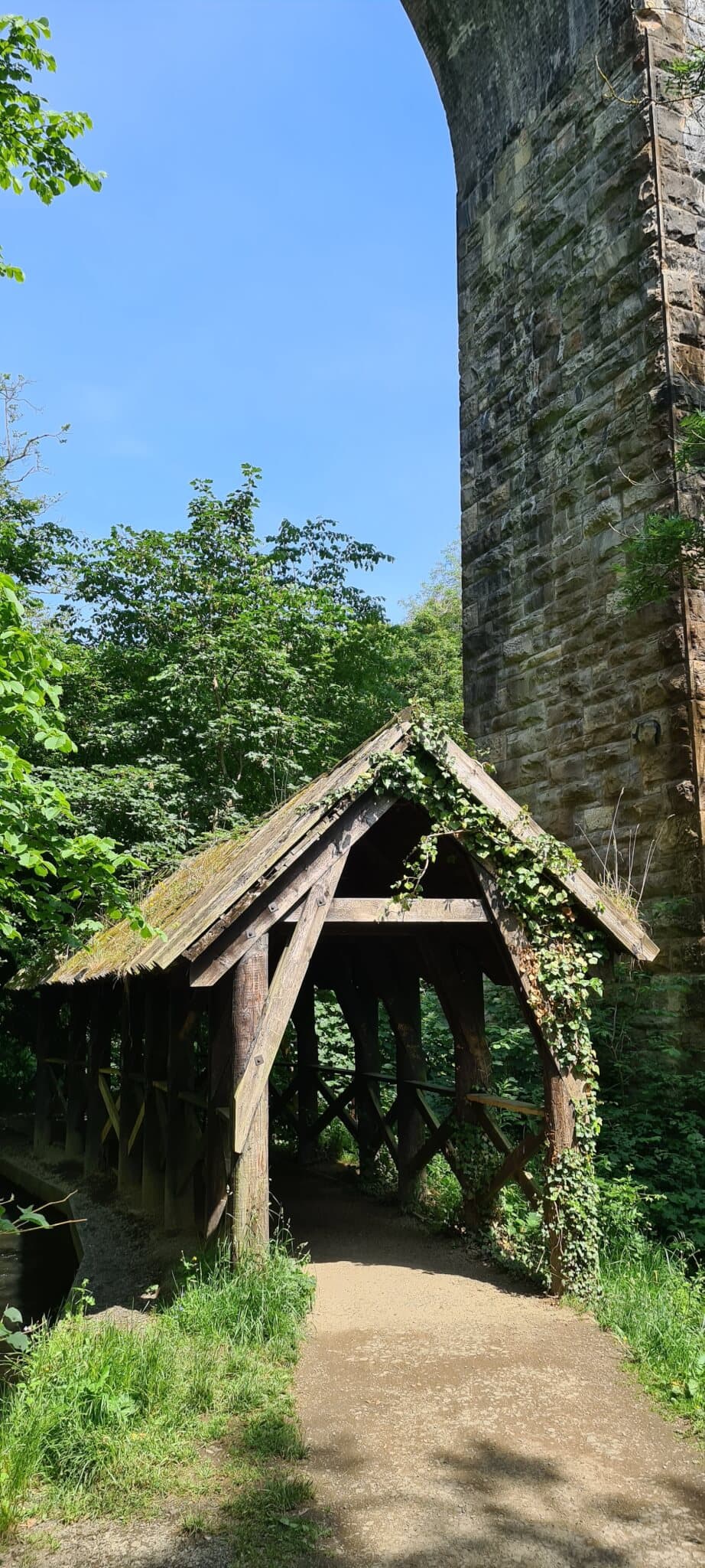 Wooden hut with a slanted roof and ivy, set below a tall stone bridge, surrounded by lush greenery under a clear sky. - Home Instead