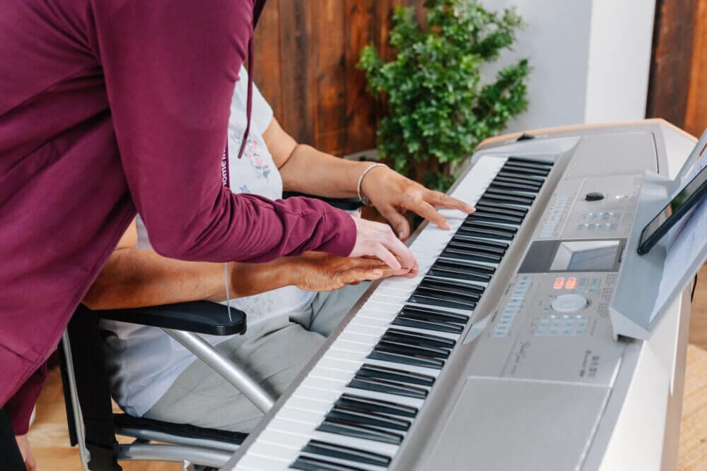 Person helps another learn piano, guiding their hands on the keys. - Home Instead