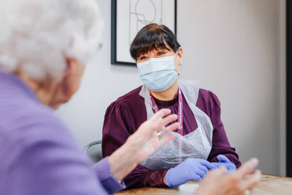A healthcare worker in PPE talks to an older woman indoors. - Home Instead