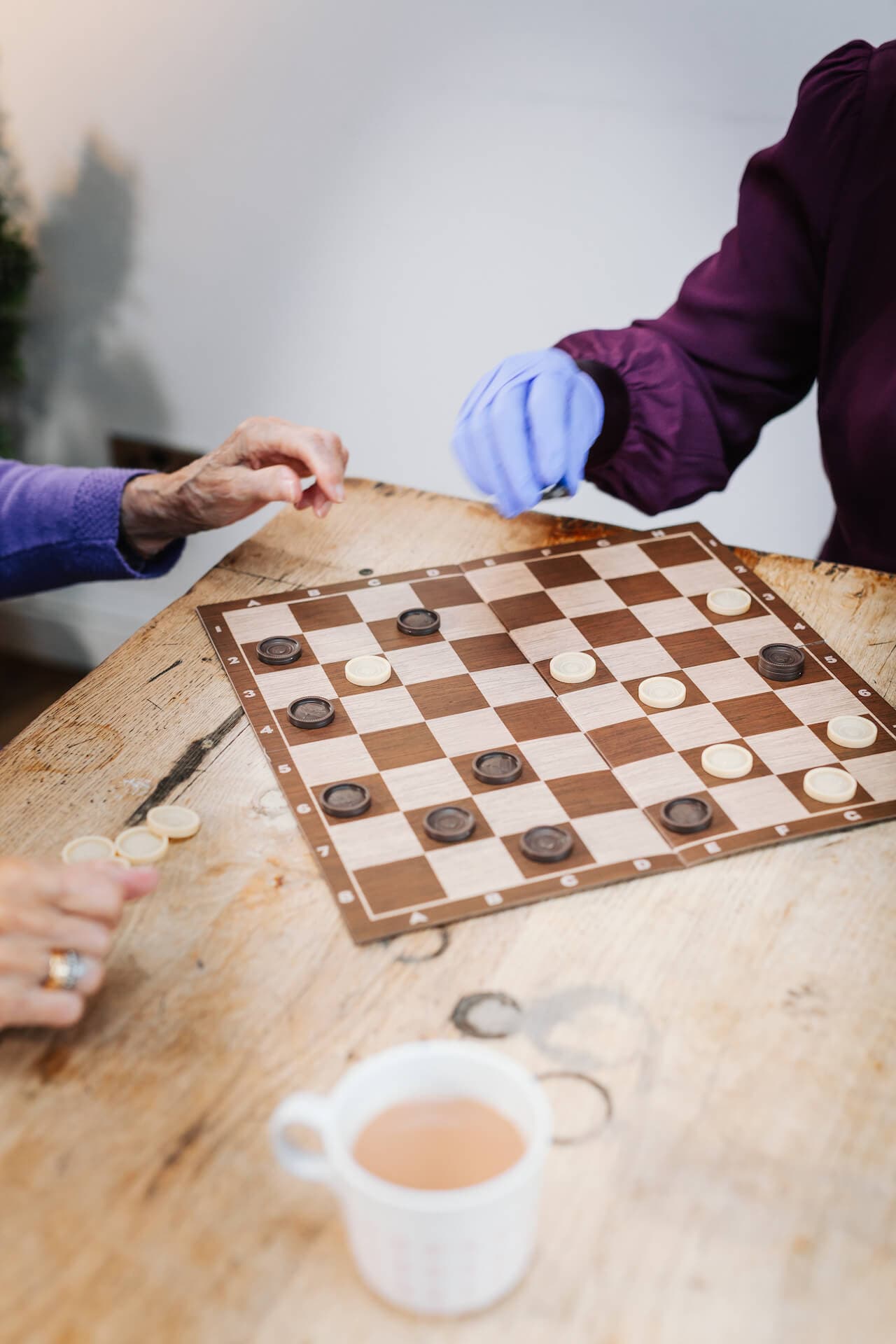 Two people playing a game of checkers on a wooden table with a cup of tea nearby. - Home Instead