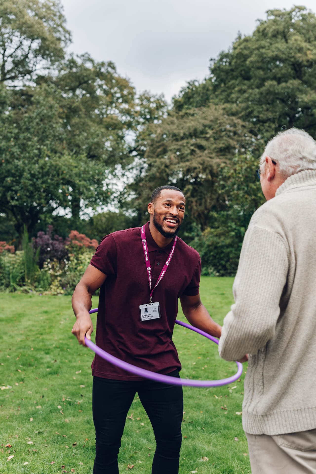 A young man with a badge and lanyard plays with a purple hula hoop outside with an older man in a sweater. - Home Instead