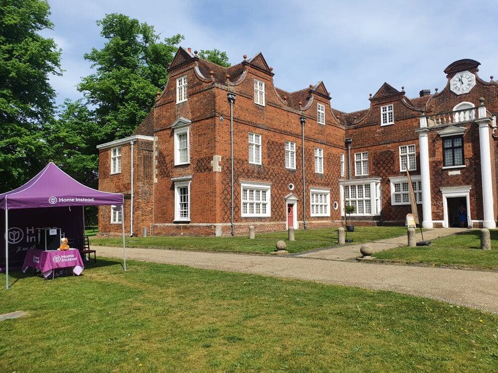 A large brick building with dormer windows next to a purple tent labeled "Home Instead" on a sunny day. - Home Instead