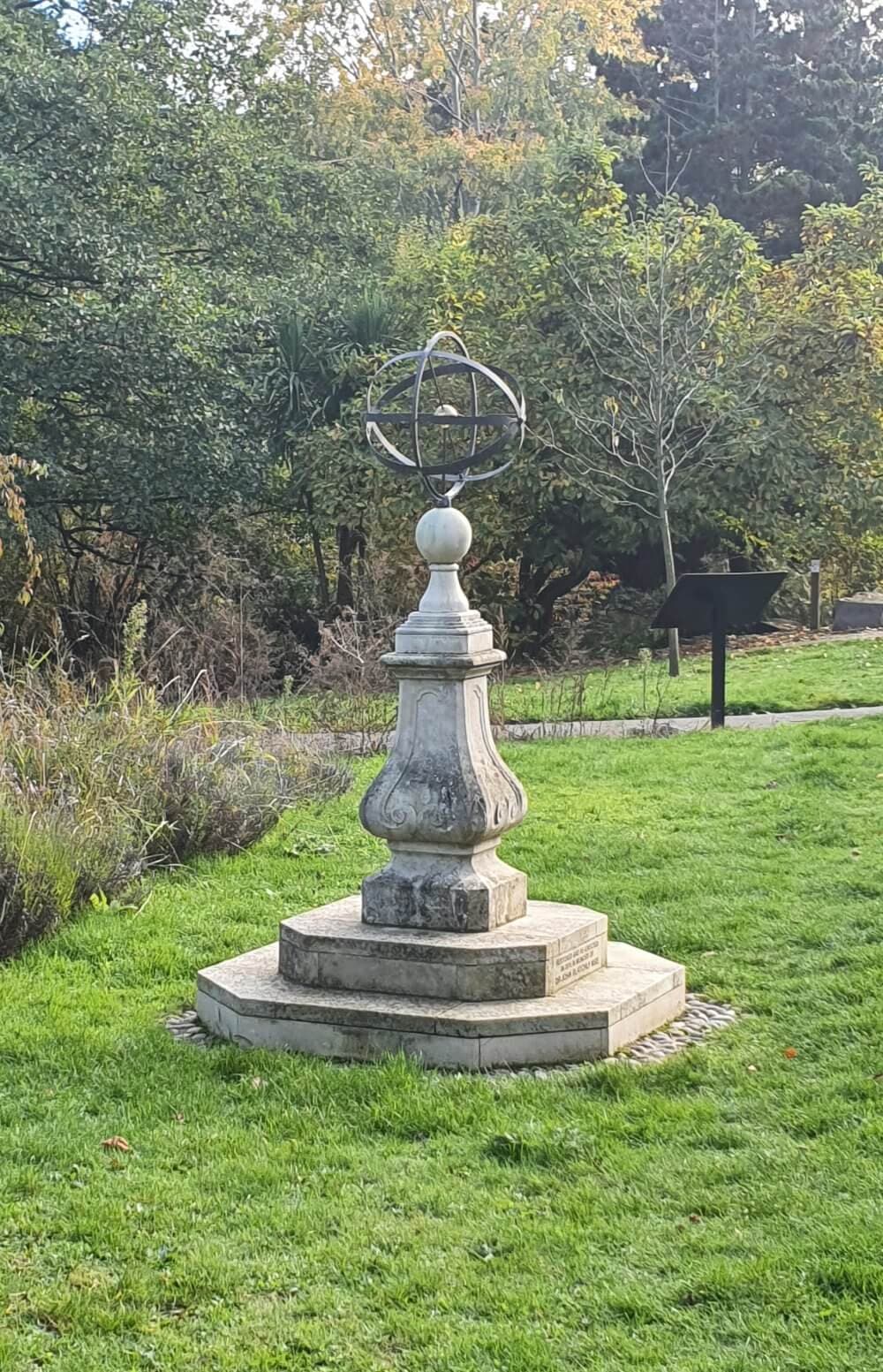 A stone sundial with a spherical armillary sphere on top, located in a green grassy park with trees in the background. - Home Instead