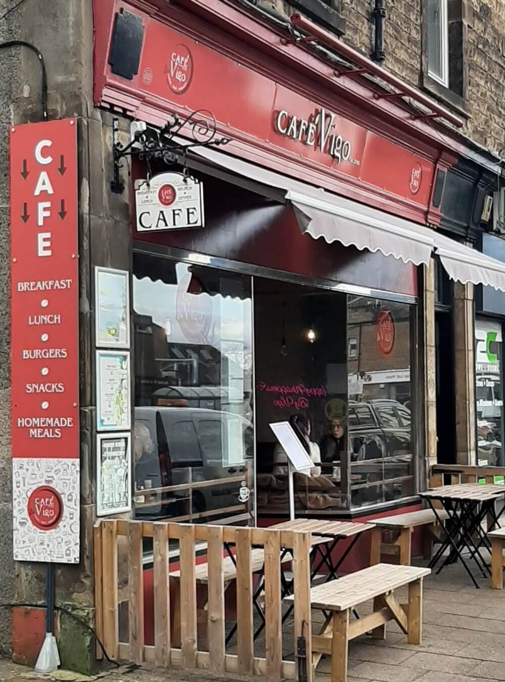 Street view of Cafe Vigo exterior with signs and a seating area, red awning, and two people seated inside by the window. - Home Instead