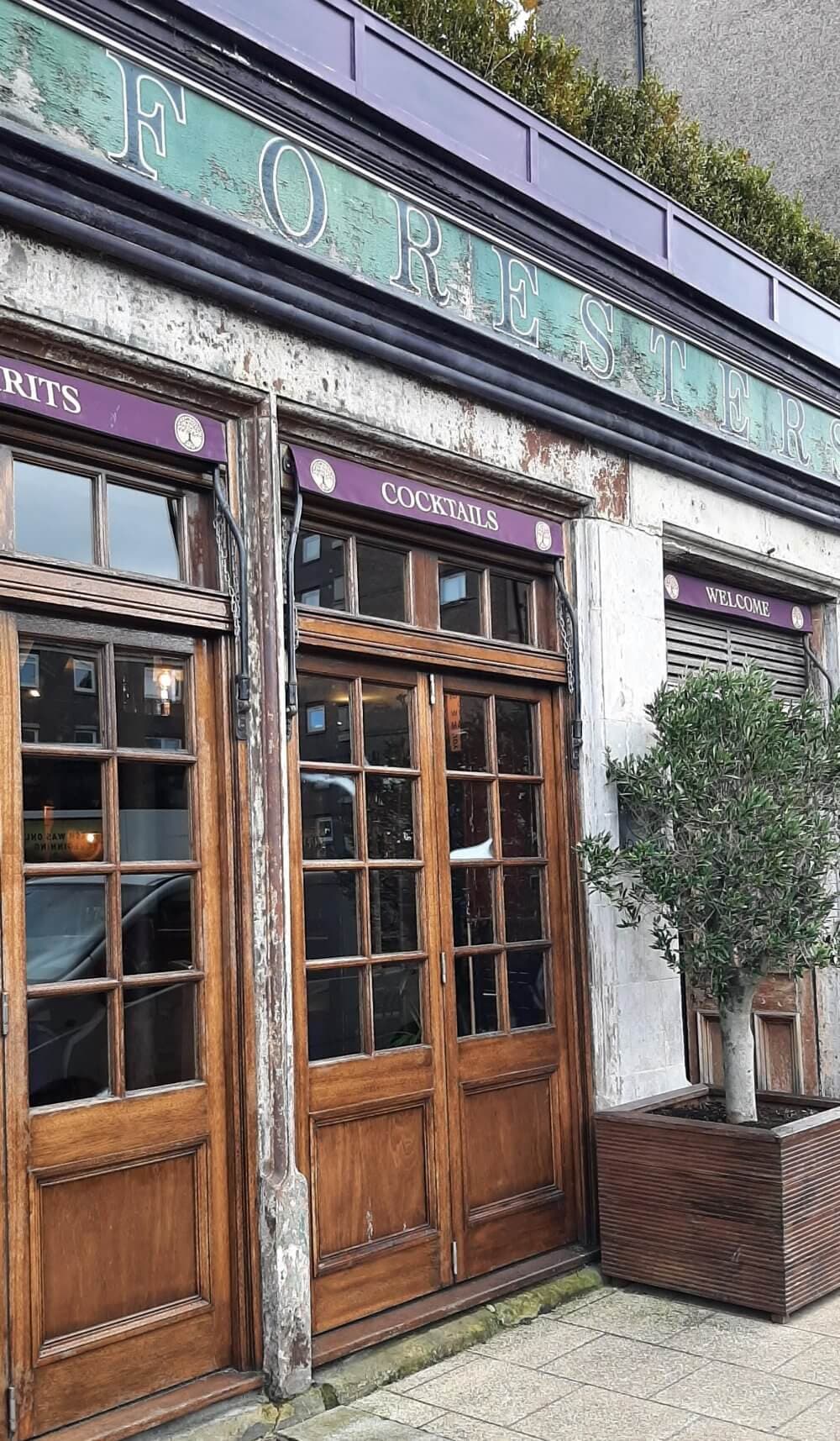 Front exterior of a rustic bar with large wooden doors, a weathered "FORESTER" sign, and a potted tree to the right. - Home Instead