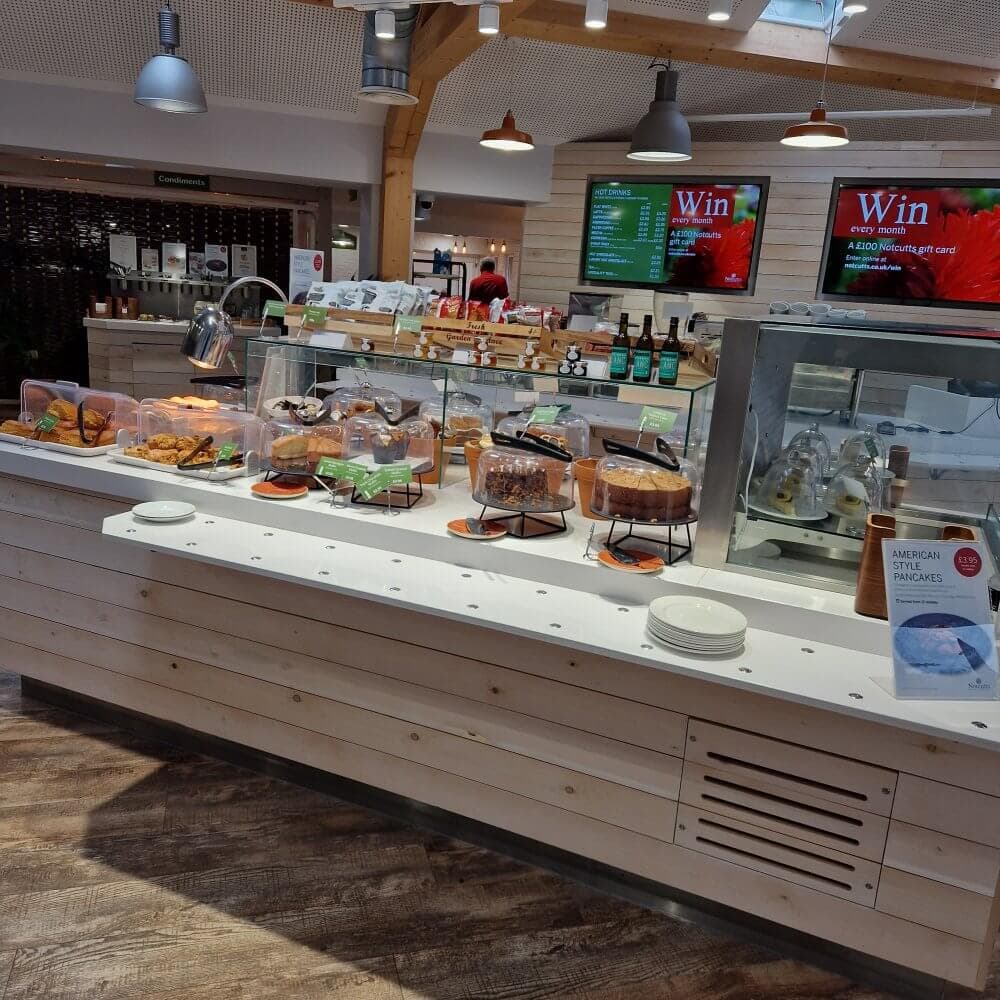 Bright cafe counter with assorted pastries, bagels, and a digital menu display. Wooden interior with pendant lights above. - Home Instead