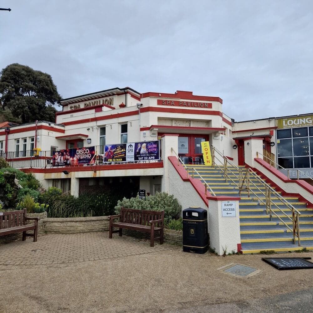 A building labeled "SPA Pavilion" with posters, a set of stairs, and a path with benches and plants in front. - Home Instead