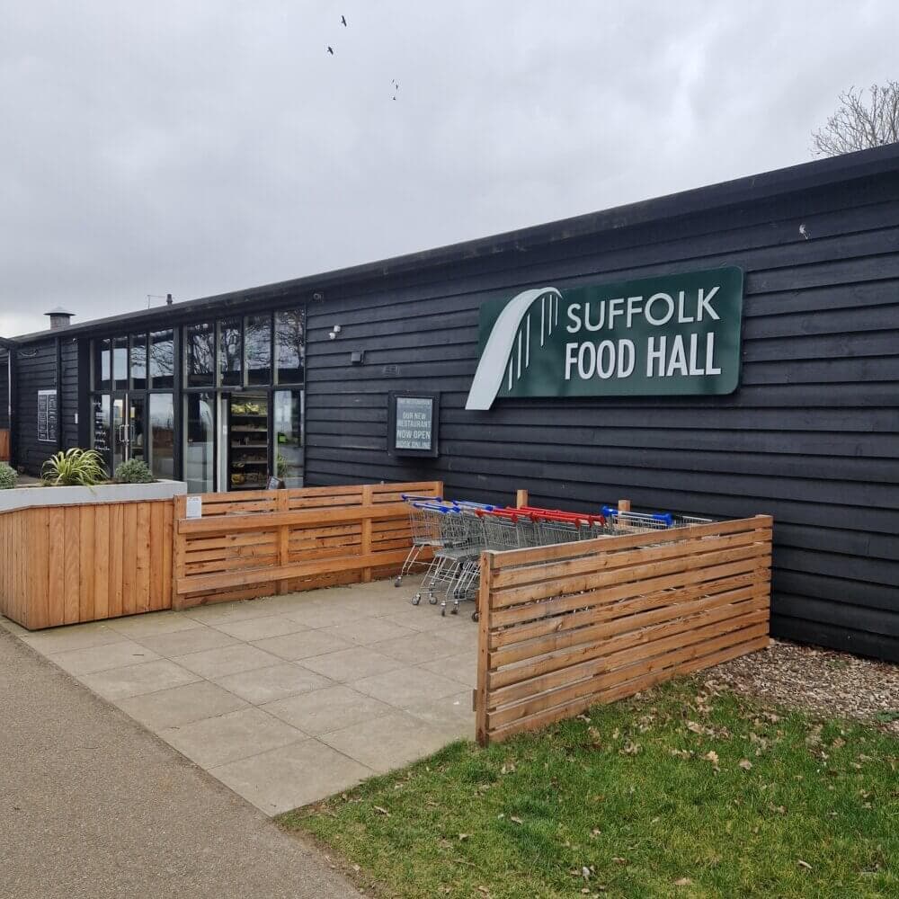 Entrance to Suffolk Food Hall with a wooden fence, shopping carts, and a sign on a dark wooden building. - Home Instead