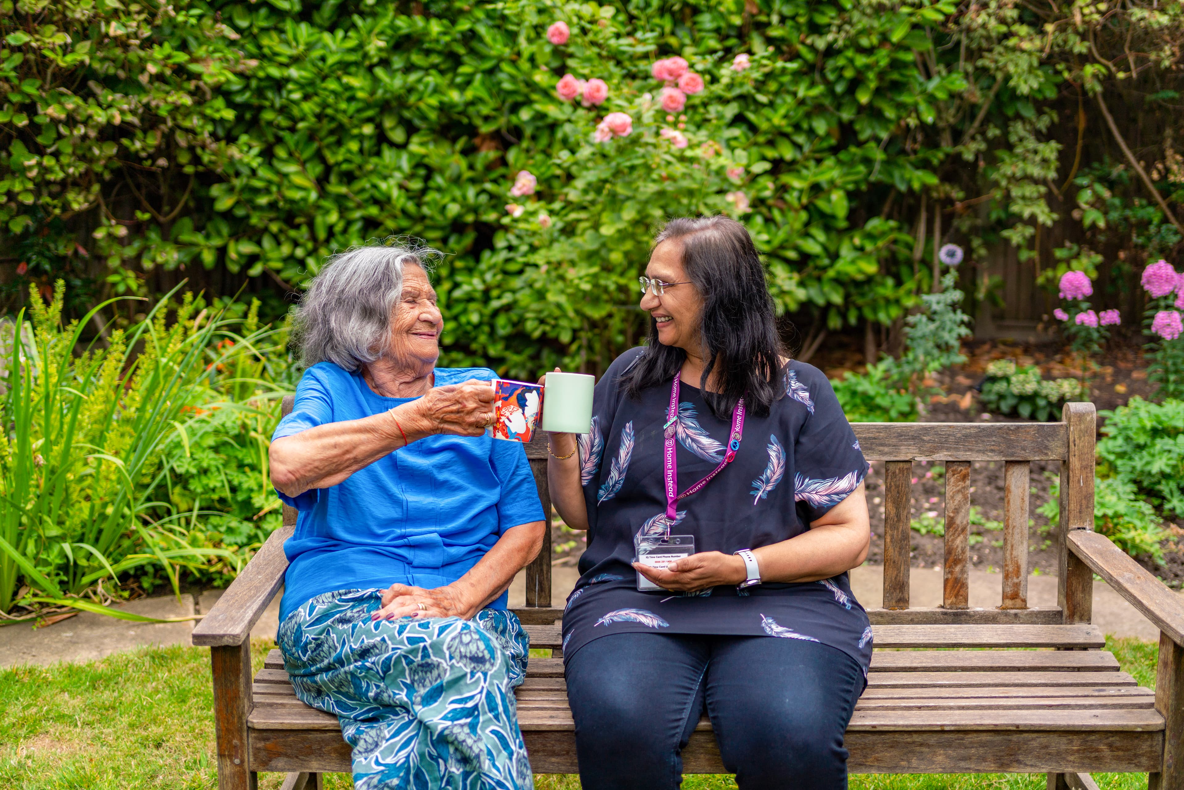 Two women smiling and toasting with coffee mugs, sitting on a bench in a garden with lush greenery and flowers. - Home Instead