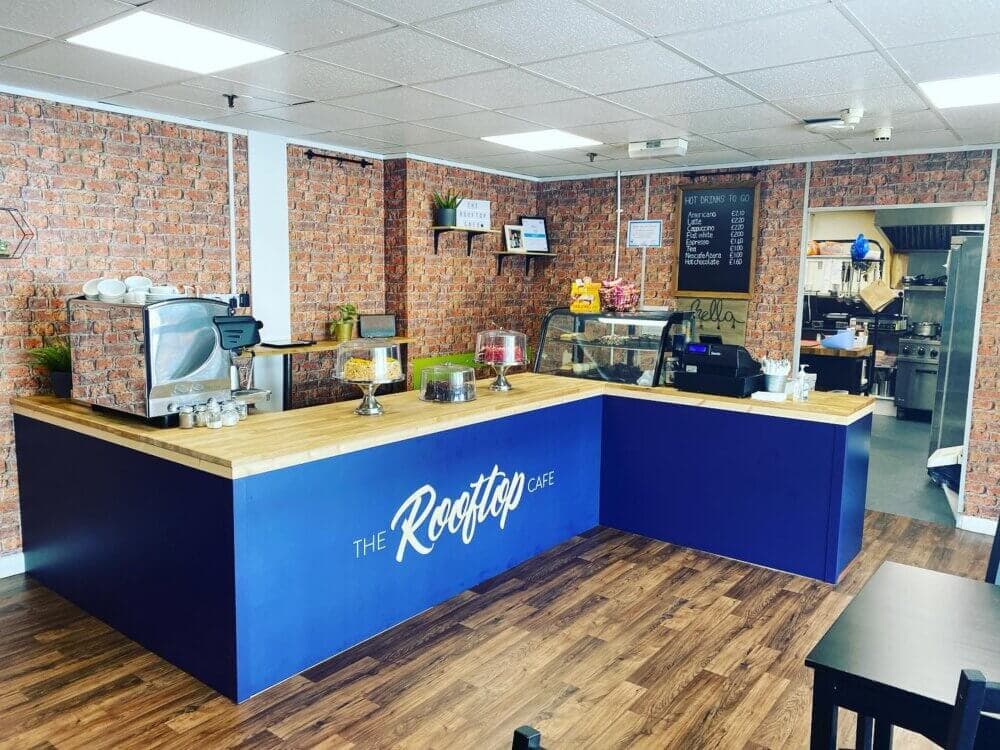 A cozy cafe with brick walls, wooden floors, a blue counter, and various items on display such as cakes and a coffee machine. - Home Instead