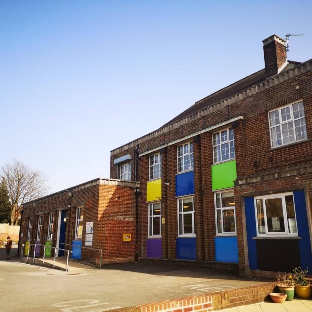 A brick school building with colorful panels below the windows and a playground in front, under a clear blue sky. - Home Instead