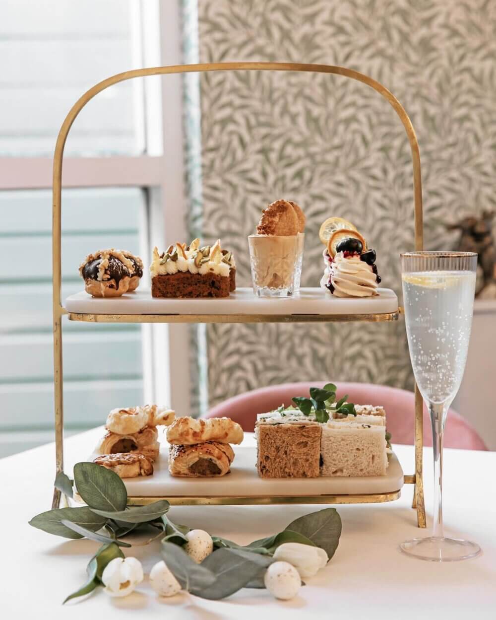 A two-tiered tray with various pastries and sandwiches next to a glass of a beverage garnished with a lemon slice. - Home Instead