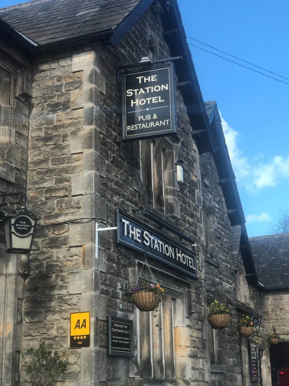 Old stone building with multiple signs saying "The Station Hotel Pub & Restaurant." Hanging baskets decorate the exterior. - Home Instead