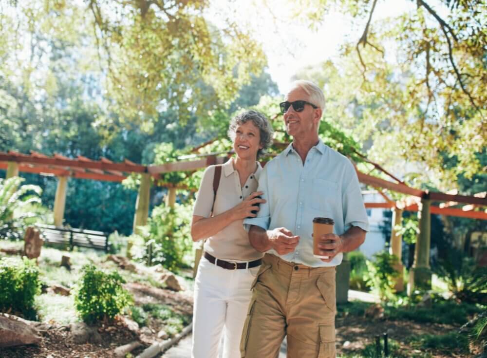 A happy elderly couple walking in a park on a sunny day, holding coffee cups and smiling. - Home Instead