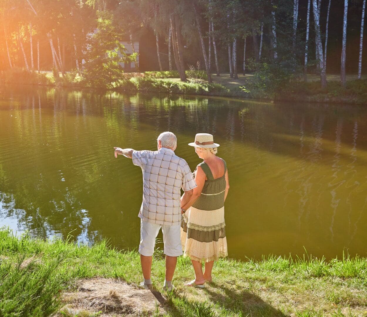 An elderly couple stands beside a lake, with the man pointing towards something while the woman looks on. - Home Instead
