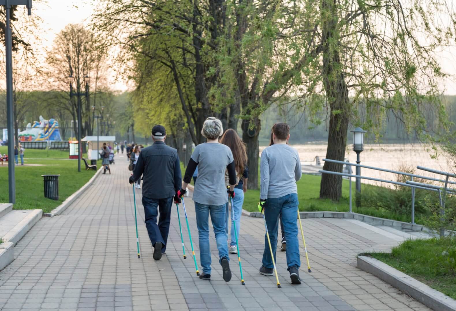 People walking with trekking poles on a paved path by the water, surrounded by trees and greenery. - Home Instead