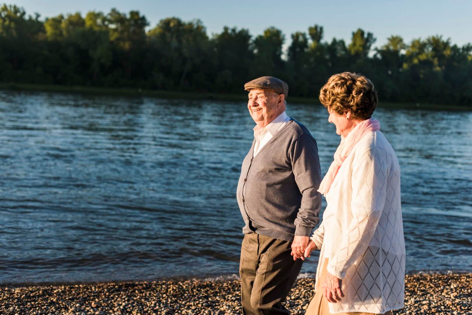 An elderly couple holding hands and walking along a beach with a body of water and trees in the background. - Home Instead