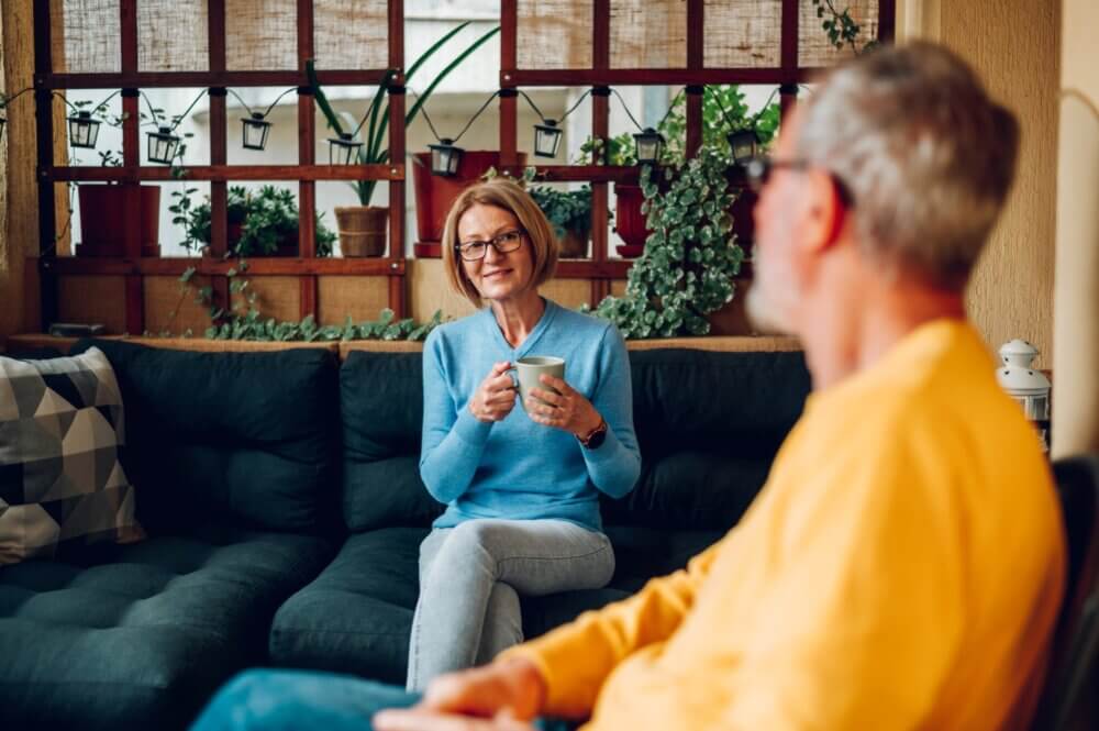 A woman with glasses holding a mug smiles at a man in glasses wearing a yellow shirt; they sit in a cozy room with plants. - Home Instead