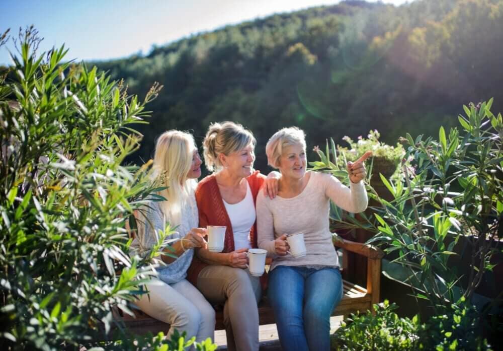 Three women sitting outdoors on a wooden bench with mugs, surrounded by greenery, one pointing into the distance. - Home Instead