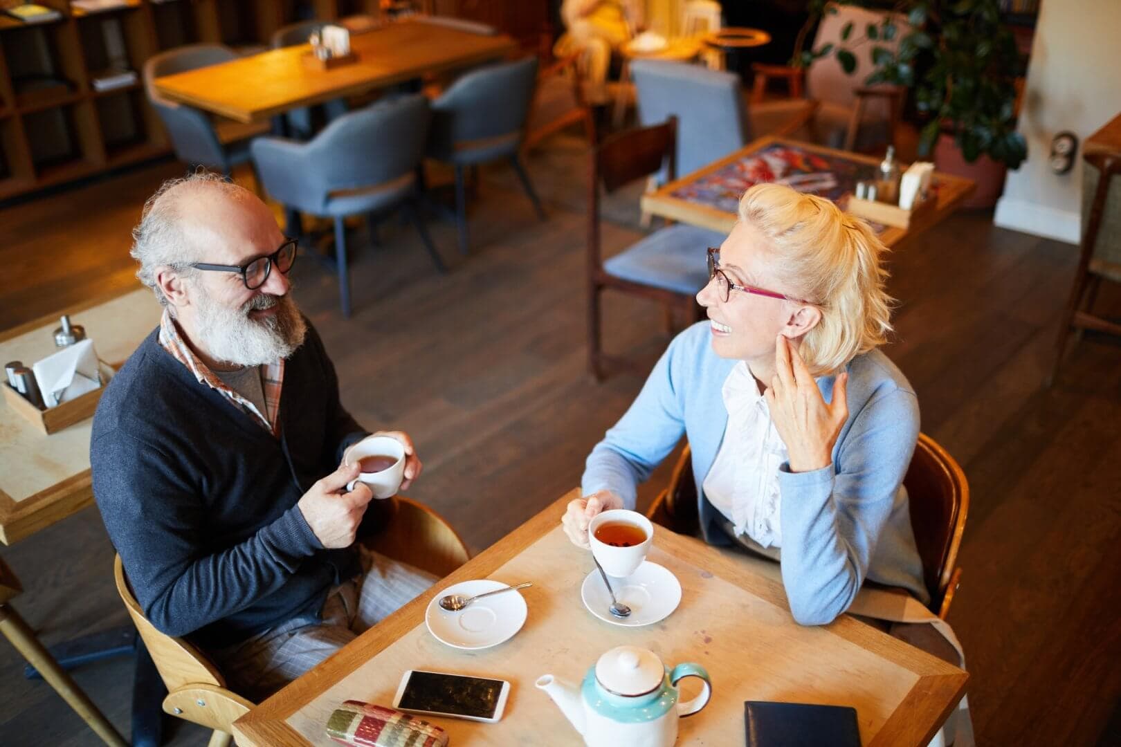 Two elderly people enjoy tea together at a cozy café, sitting at a wooden table with cups, a teapot, and a phone. - Home Instead