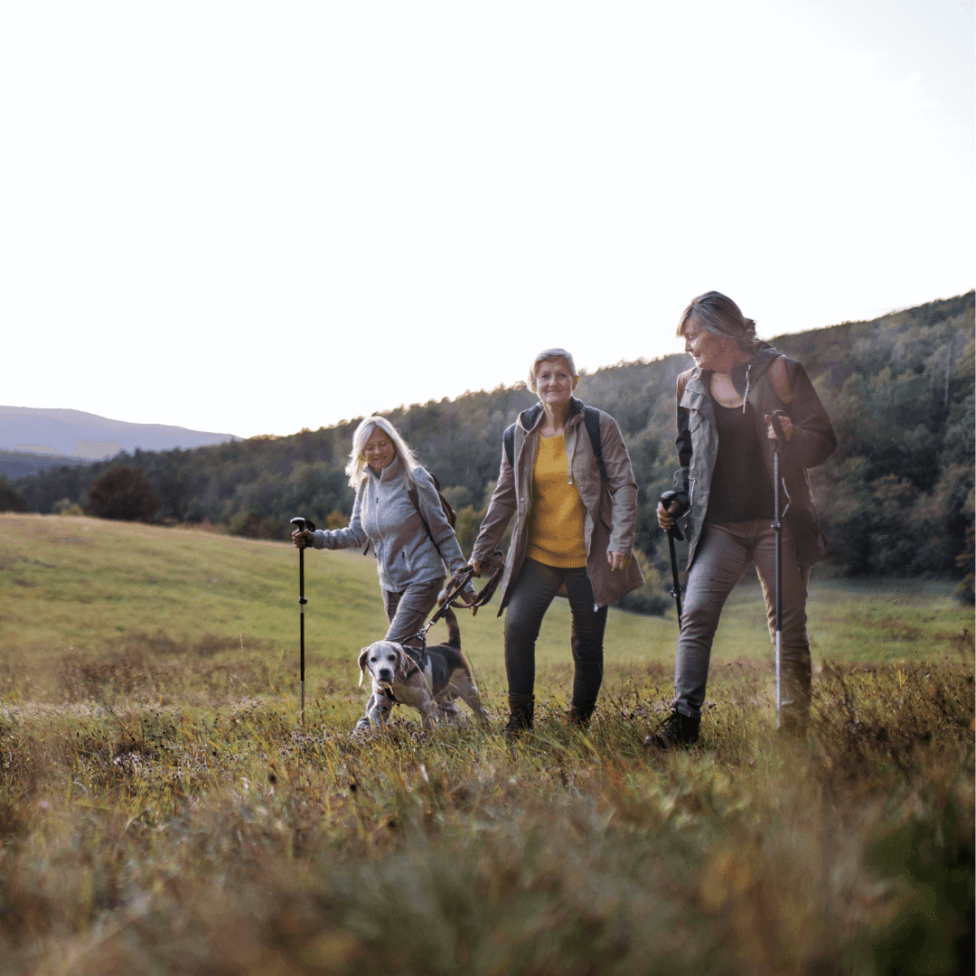 Three people hiking with walking poles and a dog on a grassy trail in a hilly, wooded area. - Home Instead