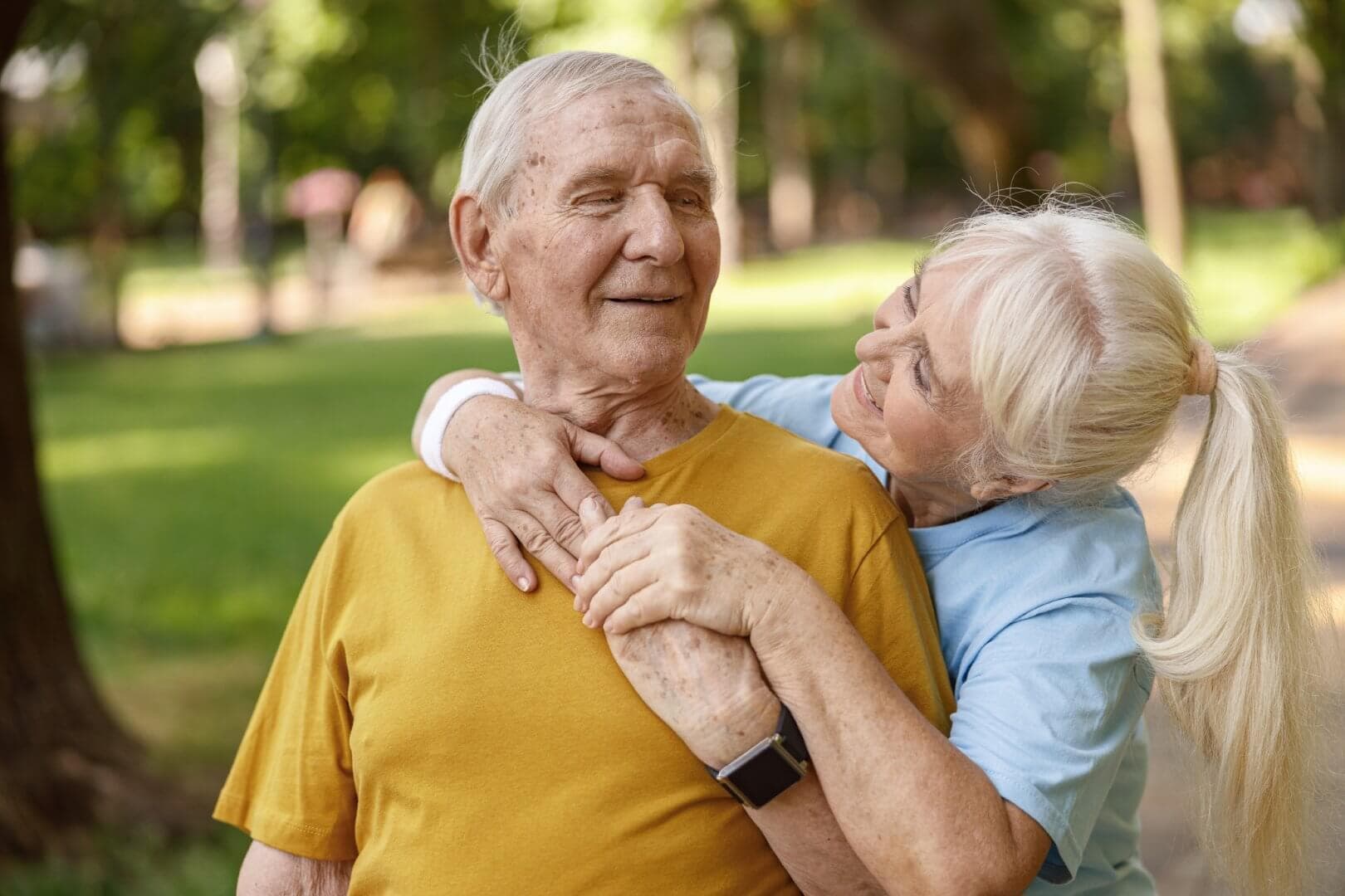 Elderly couple smiling and embracing outdoors, with the man wearing a yellow shirt and the woman a blue shirt. - Home Instead