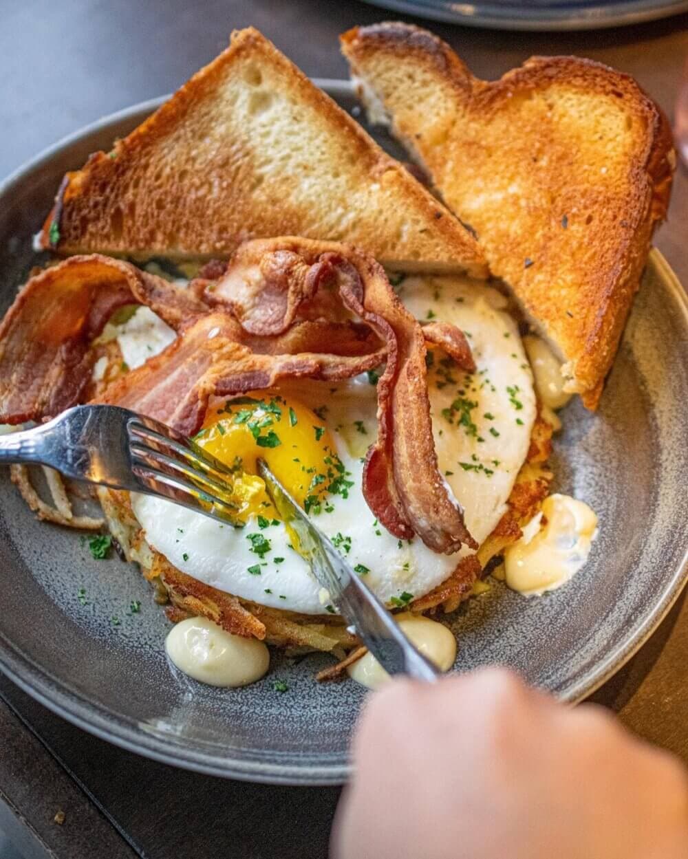 Plate with bacon, fried eggs, toast, and a hand using a fork and knife to cut into the egg yolk. - Home Instead