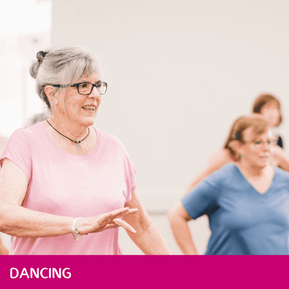 Older adults in a dance class, smiling and moving, a woman in pink shirt and glasses in foreground, text "DANCING" below. - Home Instead