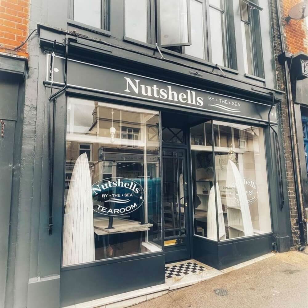 A street view of a tearoom named "Nutshells by the Sea" with surfboard decor visible inside the large front windows. - Home Instead
