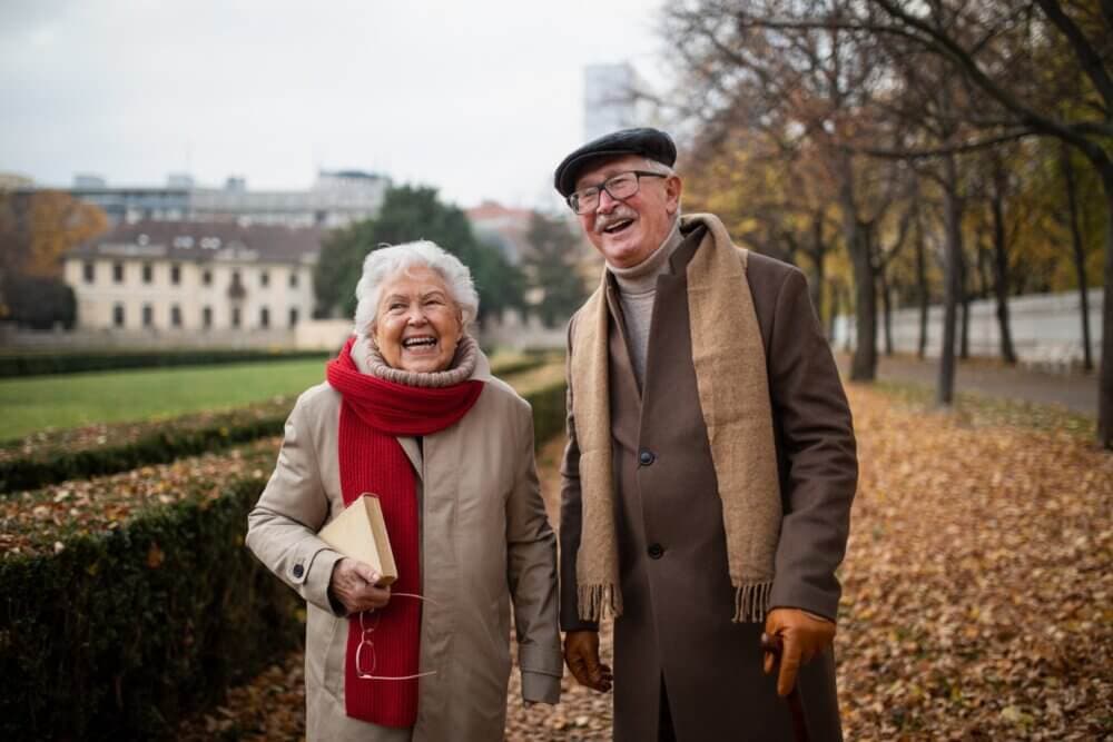 An elderly couple in coats and scarves stand on a leafy path, smiling and laughing together on an autumn day. - Home Instead