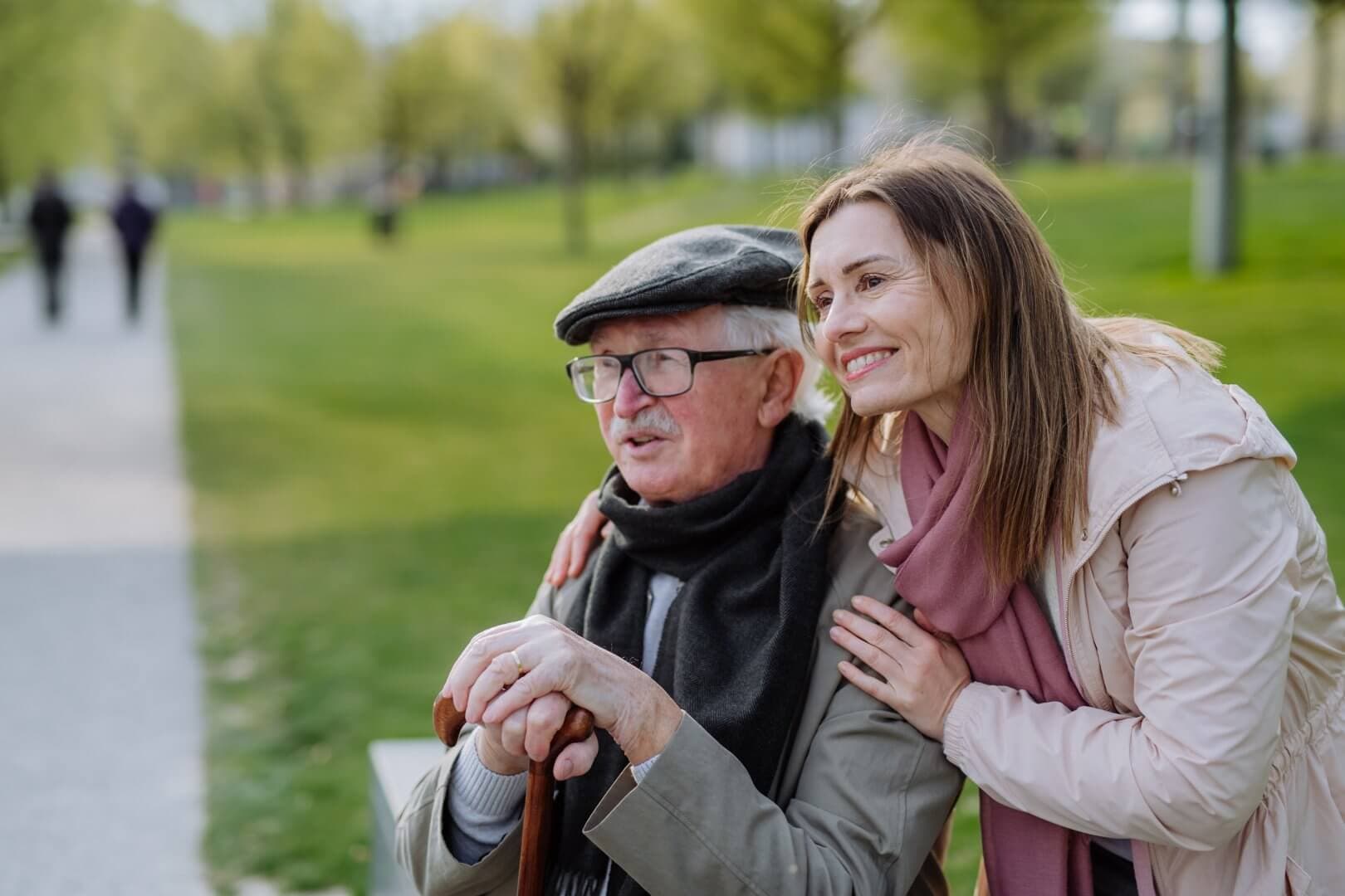 A senior man with glasses and a cane sits on a bench, while a woman stands behind him, smiling. - Home Instead