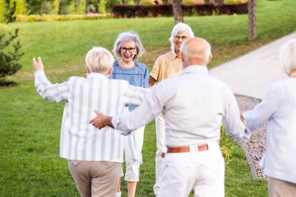 Elderly friends joyfully reuniting in a park, two with outstretched arms walking towards each other, smiling. - Home Instead
