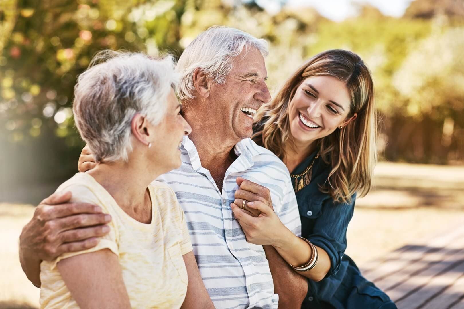 Three people smiling and embracing outdoors; two older adults and a younger adult. - Home Instead