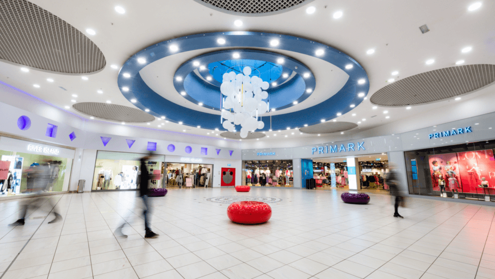 A modern shopping mall with a chandelier, vibrant decor, and various store entrances along the curved walls. - Home Instead