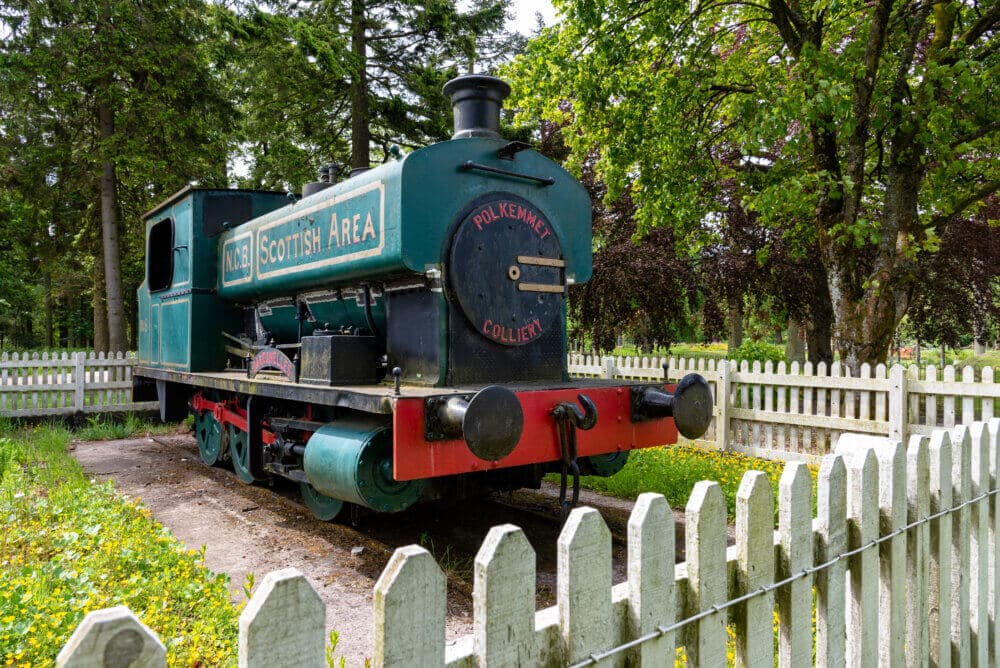 A vintage green locomotive displayed in a fenced area with trees in the background. - Home Instead