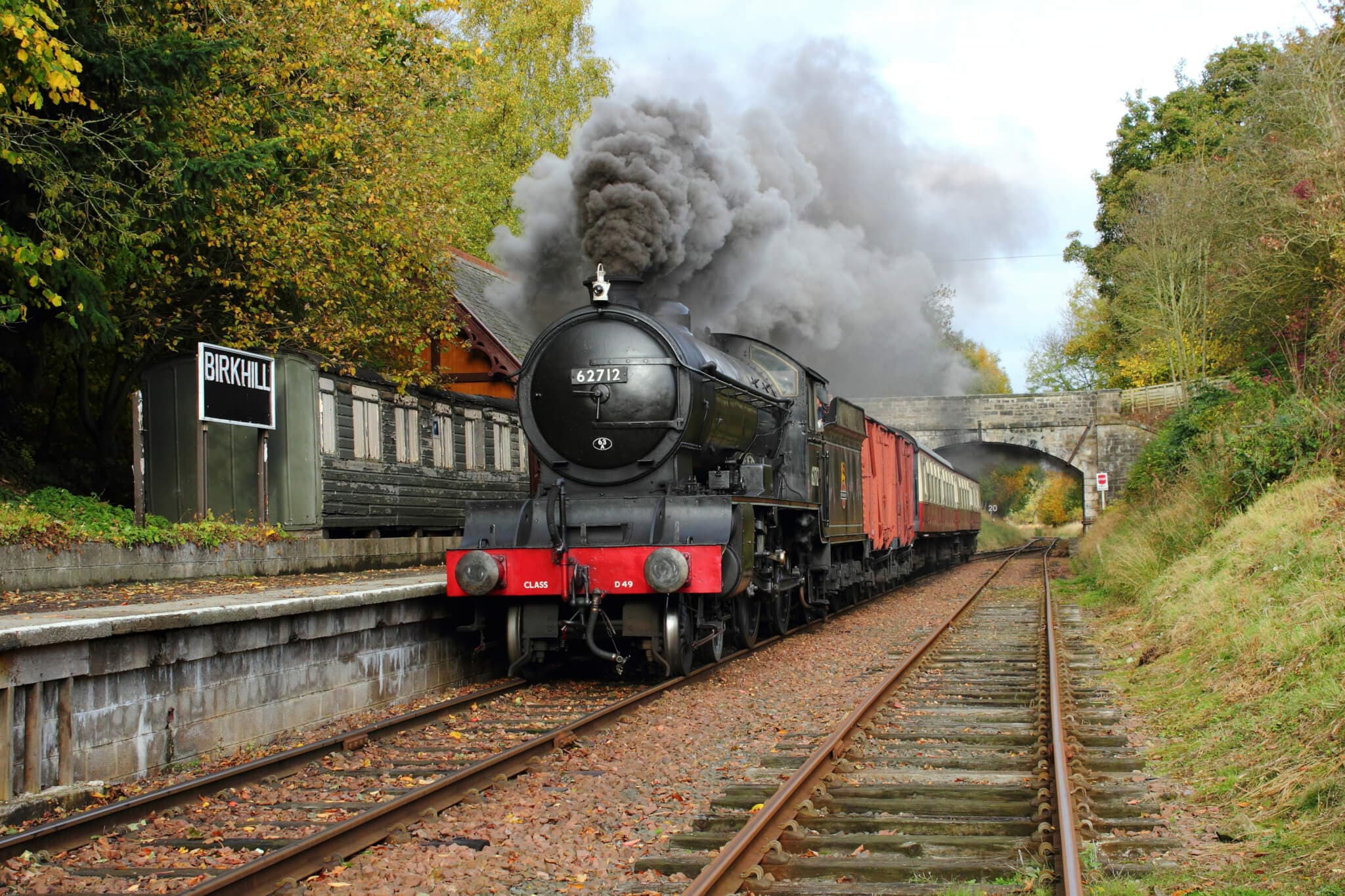 Steam train emitting smoke as it passes through Birkhill Station, surrounded by autumn trees and a stone bridge. - Home Instead