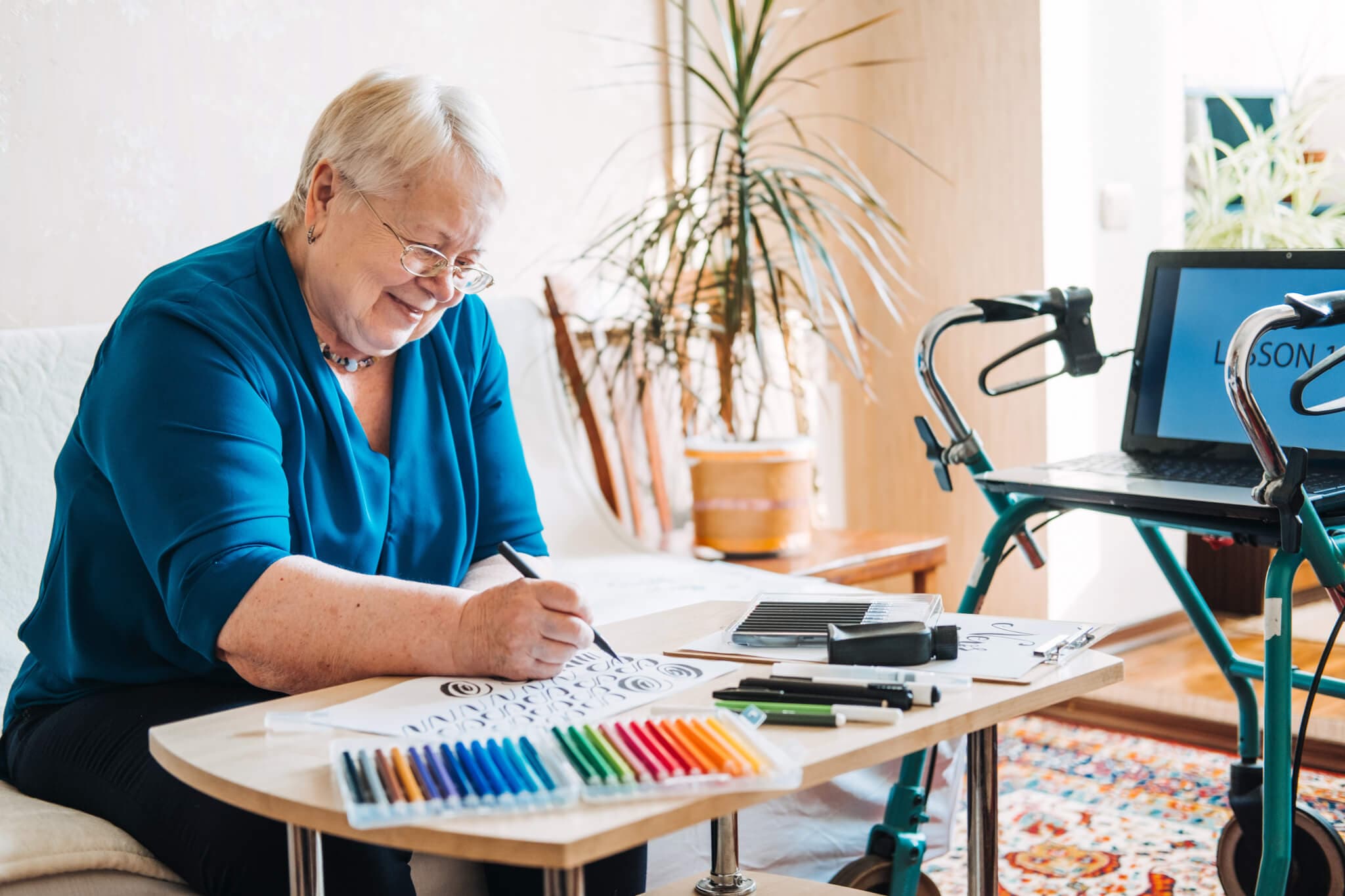 Elderly woman smiling while coloring a picture at a table, with colored markers, and walker nearby. - Home Instead