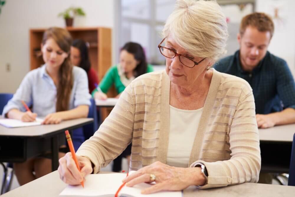 Older woman writing at a desk in a classroom with other students in the background. - Home Instead Poole