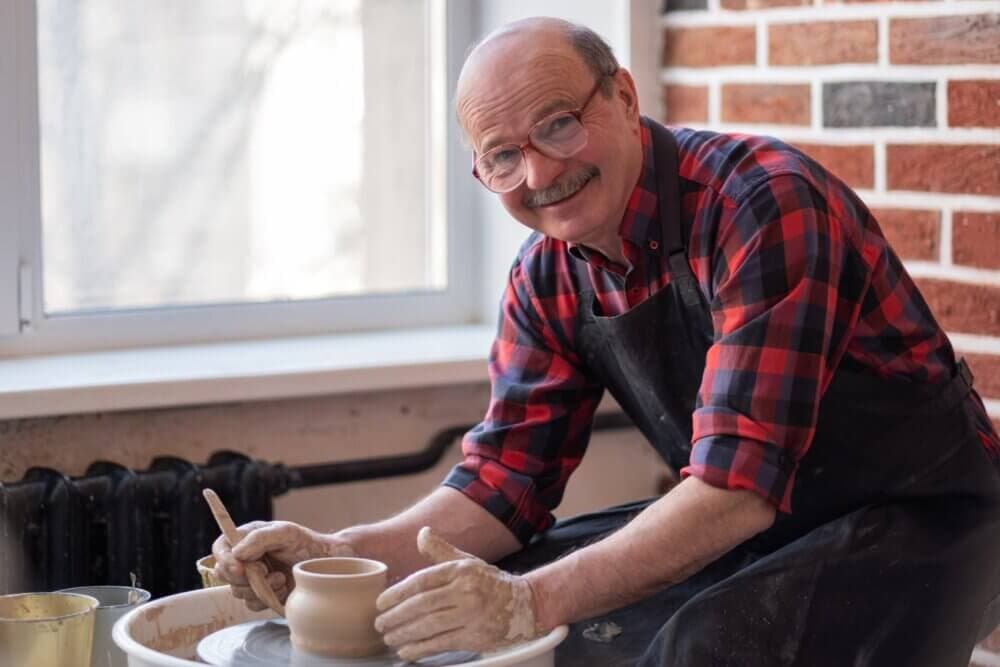 An older man wearing glasses and a plaid shirt shapes clay on a pottery wheel, smiling at the camera. - Home Instead