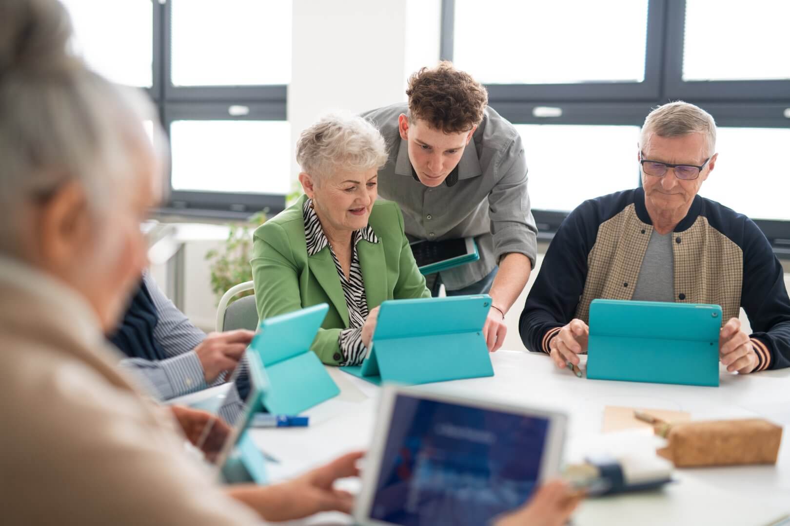 A young man assists a group of senior citizens working on tablets around a table in a bright room. - Home Instead